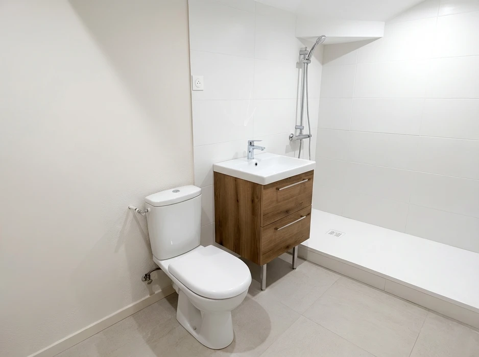 Salle de bain moderne et minimaliste avec WC à réservoir apparent, meuble vasque sur pieds en bois effet chêne avec 2 tiroirs et mitigeur chromé, receveur de douche extra-plat blanc avec colonne de douche réglable à douchette